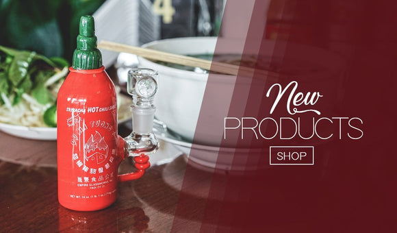 Image of a water pipe that looks like a sriracha sauce bottle, made by Empire Glassworks. The background is a table setting for an Asian style meal with a bowl of noodles and chopsticks. There is also a plate of salad greens. navigation link to new items.